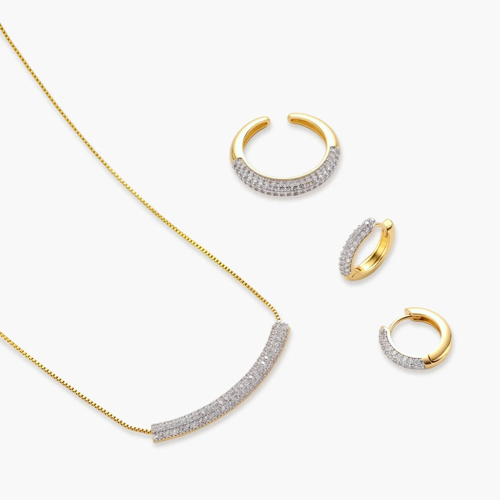 Gold-plated curved necklace, hoop earrings, and adjustable ring with pavé cubic zirconia stones on cream background.
